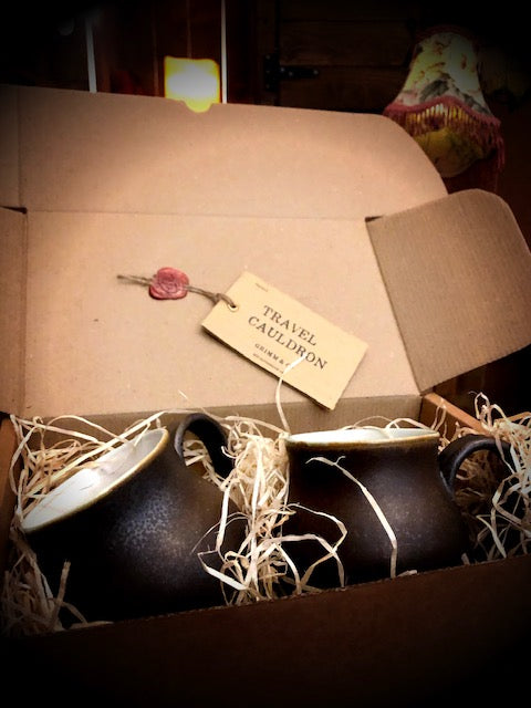 Image of two Travel Cauldrons, otherwise known as matte black ceramic mugs, nestled in wood wool in a cardboard box. A Travel Cauldron label is wax sealed to the inner box lid.