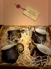 Load image into Gallery viewer, Close up image of a set of four Travel Cauldrons,  otherwise known as matte black ceramic mugs, nestled in wood wool in a cardboard box. A Travel Cauldron label is wax sealed to the inner lid of the box.