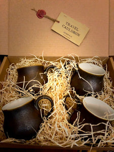 Close up image of a set of four Travel Cauldrons,  otherwise known as matte black ceramic mugs, nestled in wood wool in a cardboard box. A Travel Cauldron label is wax sealed to the inner lid of the box.
