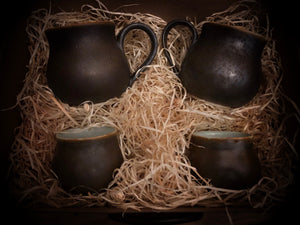 Close up, top down image of a set of four Travel Cauldrons, otherwise known as matte black ceramic mugs, nestled in wood wool. Two of the Travel Cauldrons are the standard mug size, and two are the mini espresso cup versions with no handles. 