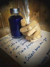 Load image into Gallery viewer, A close up image of a Starlight ink bottle next to a wooden mannequin hand holding a white feather quill, with a parchment page of writing done in the blue and silver shimmering ink.