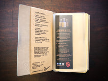 Load image into Gallery viewer, Image of a journal opened at the front, showing the clear storage sleeve containing a bookmark, and the part of the kraft label on the inner cover, which lists the faux ingredients and side effects.