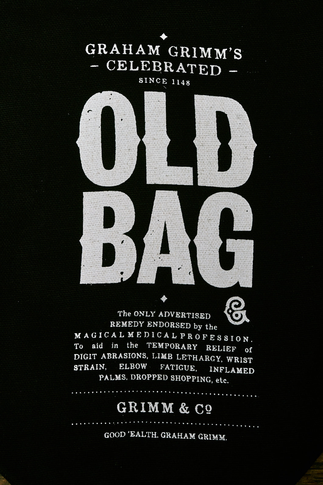 Close up image of the printed lettering of the old bag:  'Graham Grimm's Celebrated - since 1148 - OLD BAG. The only advertised remedy endorsed by the Magical Medical Profession. To aid in the temporary relief of digit abrasions, limb lethargy, wrist strain, elbow fatigue, inflamed palms, dropped shopping, etc.' Underneath that is written: 'Grimm & Co, Good 'ealth Graham Grimm'.