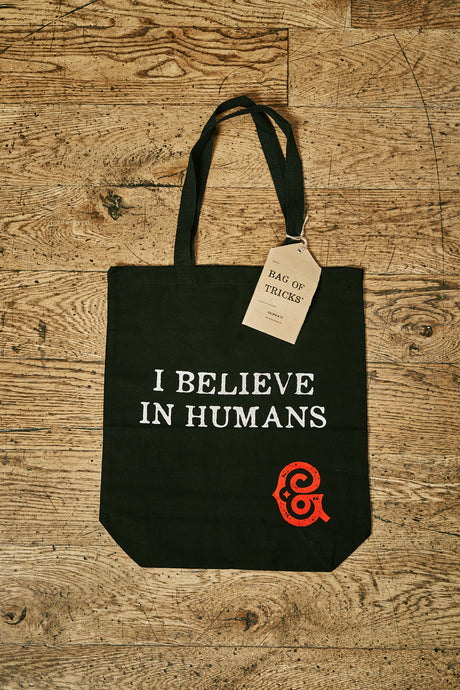 Image of black cotton tote book bag with white printed slogan on front saying 'I BELIEVE IN HUMANS ' with the red Grimm & Co 'G' monogram in the bottom right corner