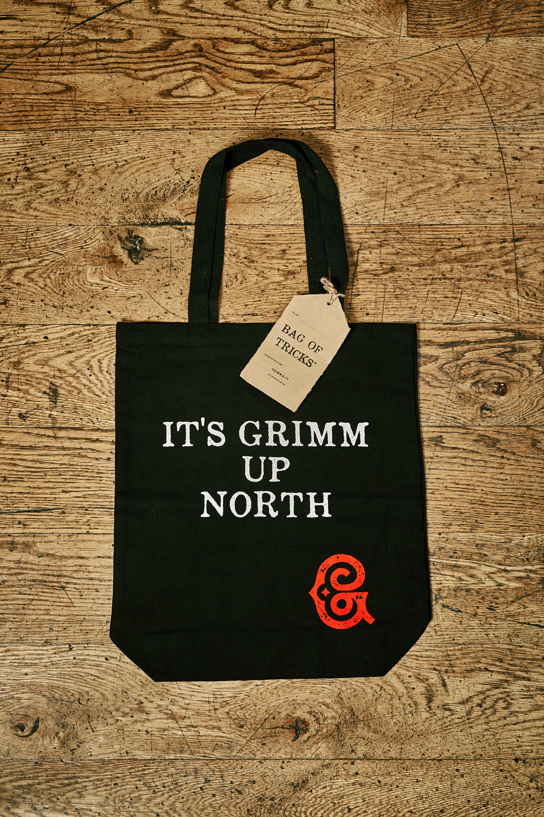 Image of black cotton tote book bag with white printed slogan on front saying 'IT'S GRIMM UP NORTH' with the red Grimm & Co 'G' monogram in the bottom right corner