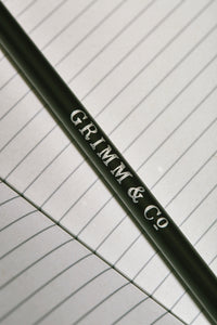 Close up details of lettering on one side of a Word Wand, otherwise known as a black pencil with a black eraser at the top. The silver lettering in the image reads 'Grimm & Co.'