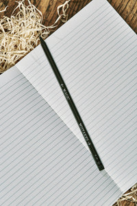 Image of a Word Wand, otherwise known as a black pencil with silver printed writing along the length and a black eraser at the top. The lettering on this side of the pencil reads 'Mistakes' with an arrow pointing towards the eraser, and 'Magic' with an arrow pointing towards the lead. The Word Wand rests on a lined kraft note book.