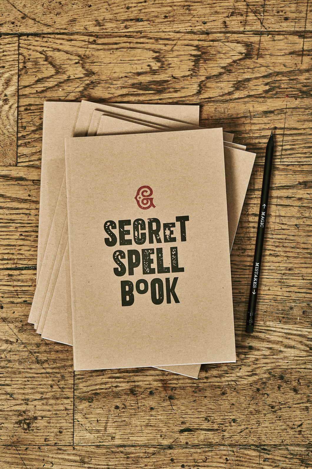 Image shows a few kraft card notebooks in a pile with the top one displaying the slogan 'SECRET SPELL BOOK'. Notebooks are shown with a Word Wand pencil.