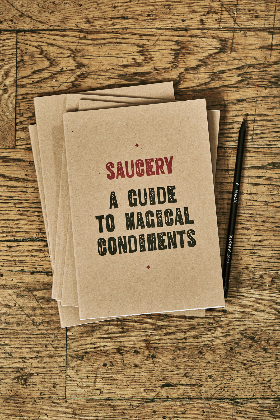 Image shows a few kraft card notebooks in a pile with the top one displaying the slogan 'SAUCERY: A GUIDE TO MAGICAL CONDIMENTS'. Notebooks are shown with a Word Wand pencil.