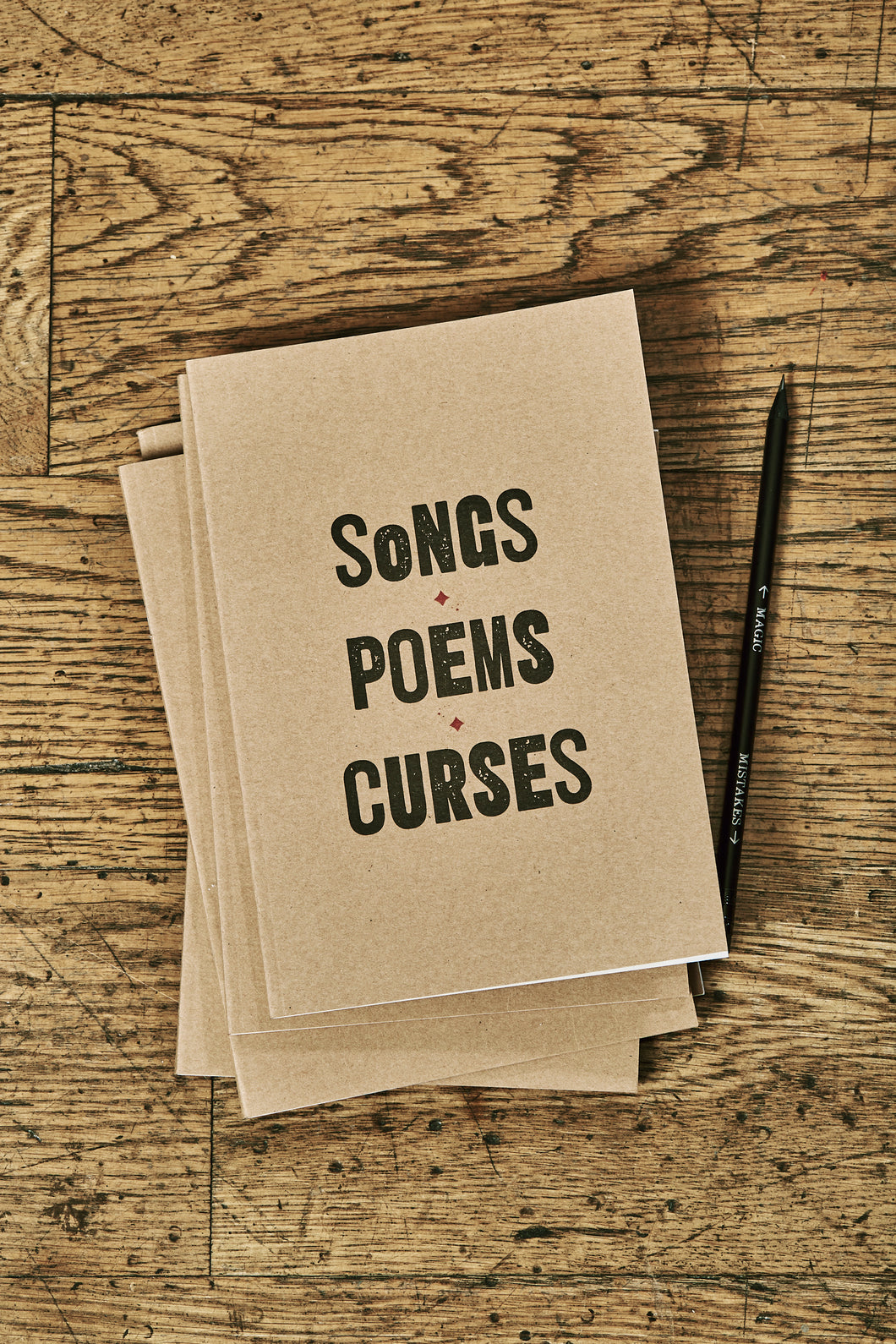 Image shows a few kraft card notebooks in a pile with the top one displaying the slogan 'SONGS, POEMS, CURSES'. Notebooks are shown with a Word Wand pencil.