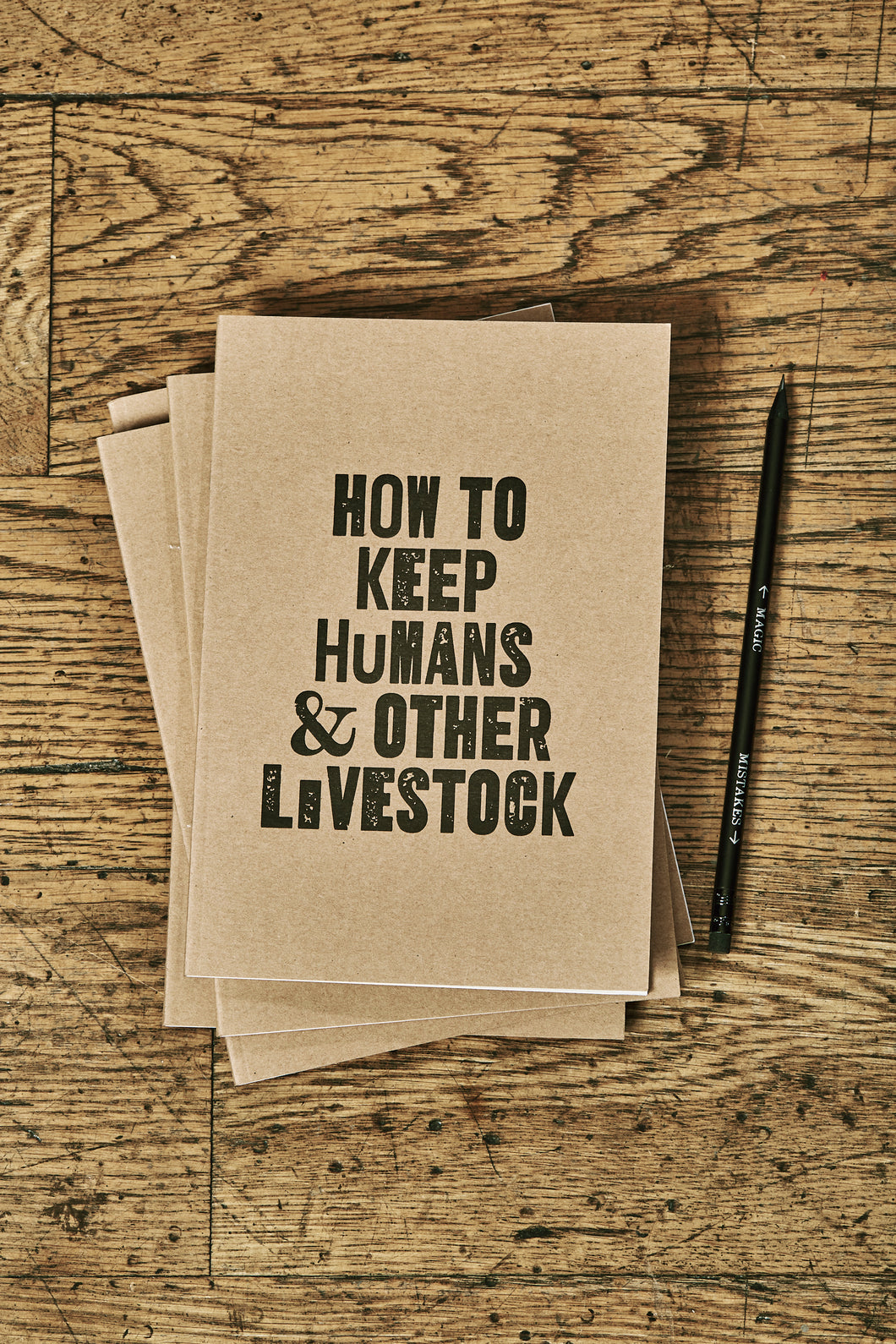 Image shows a few kraft card notebooks in a pile with the top one displaying the slogan HOW TO KEEP HUMANS & OTHER LIVESTOCK'. Notebooks are shown with a Word Wand pencil.