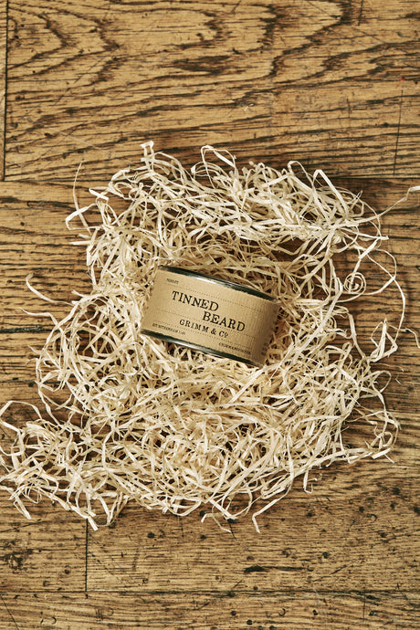 Image of a Tinned Beard, otherwise known as a 125ml tin wrapped in a brown kraft label, containing an artificial beard. Tin in image is closed and placed on its side to show the name on the label, resting on a pile of wood wool.