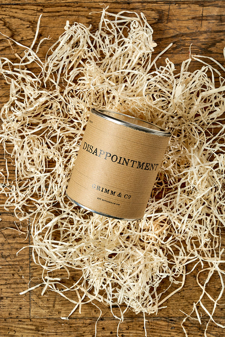 Image of a tin of Disappointment with a kraft label shown sat in a nest of wood wool. 
