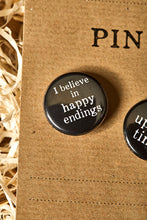 Load image into Gallery viewer, Image of a black button badge with the slogan &#39;I believe in happy endings&#39; in white text.