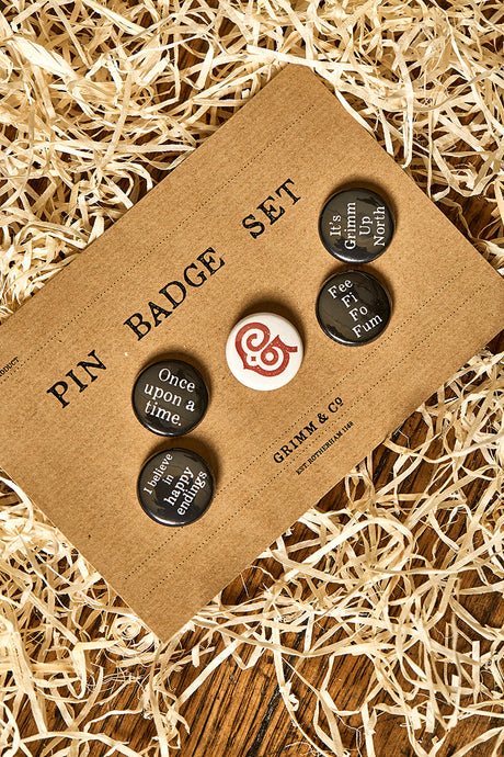 Image of set of 5 button badges on a kraft card backing. Slogans incclude Once upon a time, I believe in happy endings, Fee Fi Fo Fum, It's Grimm Up North and the red Grimm & Co 'G' monogram
