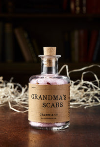 Image shows a bottle of Grandma's Scabs potion, a blass bottle with cork containing pale pink bath salts and dried rose petals. Bottle is wrapped with a kraft paper label.