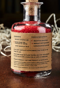 Image shows a bottle of Middle Aged Vitriol, otherwise known as red, scented bath salts in a glass bottle with cork lid and a kraft paper label. Detail of back of label showing faux ingredients and side effects for mortals.