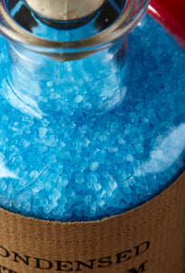 Close up image of Condensed Enthusiasm, otherwise known as scented, baby blue bath salts in a glass bottle with cork