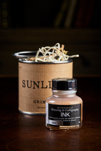 Image of a tin of Sunlight, otherwise known as a gold drawing or writing ink. A bottle of the ink stands in front of the tin. The tin is wrapped with a kraft paper label and has the lid slightly removed, showing wood wool inside.