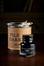 Load image into Gallery viewer, Image of a tin of Vile Darkness, otherwise known as a black drawing or writing ink. A bottle of the ink stands in front of the tin. The tin is wrapped with a kraft paper label and has the lid slightly removed, showing wood wool inside.