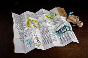 A detail image of the unfolded illustrated story written by Sir Bob Geldof and illustrated by Lewis James. Next to story is the hessian pouch of glow in the dark pebbles and tiny jar of glitter.