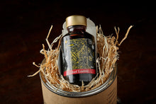 Load image into Gallery viewer, Image of Berry Sparkle, a red and gold shimmer dipping ink. Bottle of ink shown propped out of its box in bed of wood wool