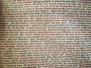 Detail of story excerpt scarf featuring text from Hamlet in red cursive font on white background.