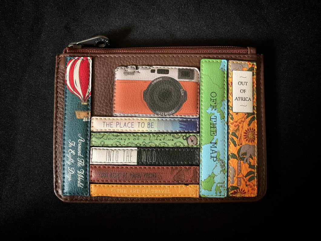 Image shows a brown Yoshi leather zip coin purse featuring an appliqued leather design of travel related classic book spines and a leather camera sat on the books.