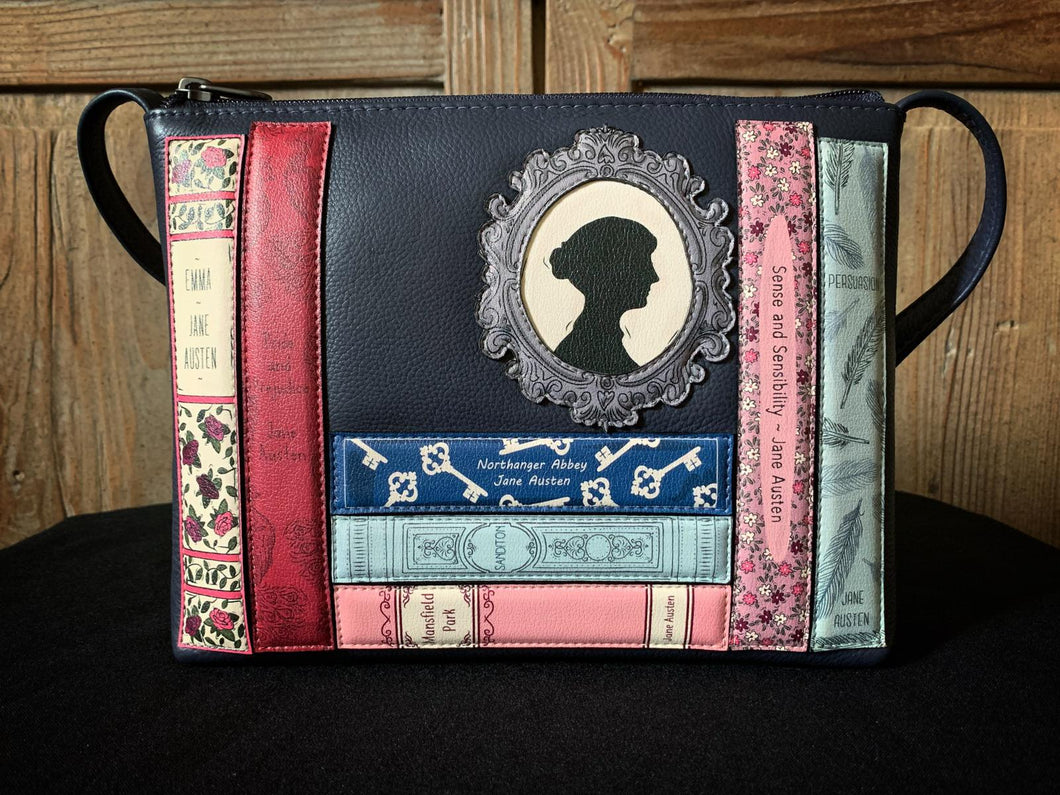 Image of a Yoshi navy blue leather cross-body handbag, with design showing appliqued book spines across the front with classic titles from Jane Austen, including Pride & Prejudice, Northhanger Abbey, Sense & Sensibility, and Emma. Also features an appliqued cameo resting above the books.
