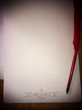 Load image into Gallery viewer, Detail of the cream parchment style paper with printed wording saying &#39;Changing lives one story at a time&#39; and the red biro quill laid next to it