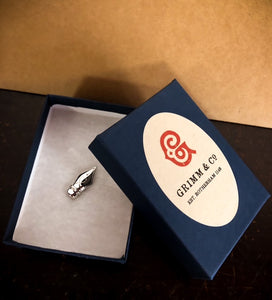 Image of the fountain pen nib lapel pin sat inside a small navy blue gift box with a Grimm & Co label. There is a layer of sponge for the pin to sit on.