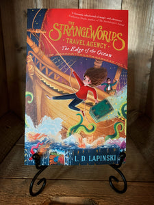 Image of the front cover of the paperback book The Strangeworlds Travel Agency: The Edge of the Ocean, written by L.D. Lapinski. Displayed on a book stand .