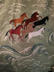 Image of the story extract scarf 'Extract from a tale on the rugged plains' with a printed pattern of linear rolling hills in white and dark teal with groups of brown and tan horses galloping amid a linear white dust cloud on a background of olive green. Image shows detail of full printed pattern.