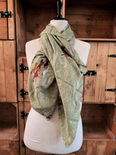 Load image into Gallery viewer, Image of the story extract scarf &#39;Extract from a tale on the rugged plains&#39; with a printed pattern of linear rolling hills in white and dark teal with groups of brown and tan horses galloping amid a linear white dust cloud on a background of olive green. Scarf displayed tied around a mannequin.