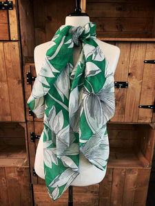 Image of the story extract scarf 'Extract from a tale in the jungle (green leaves)' with a printed pattern of large tropical stems with white tropical and leafy flowers with leaves on a bright green background. Scarf displayed tied around a mannequin.