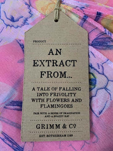 Image of the story extract scarf 'Extract from a tale in a land of wonder' with a printed pattern of yellow, pale pink and white daffodils with purple stems and leaves on a baby pink background. Kraft paper label is shown with description 'An extract from a tale of falling into frivolity with flowers and flamingos'.