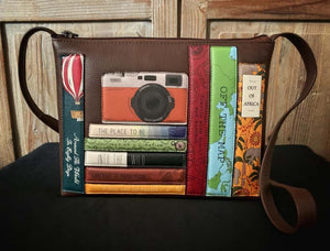 Image shows a brown Yoshi leather cross-body handbag with appliqued design featuring book spines with travel-themed titles and an orange vintage appliqued camera sat on top of the books. 