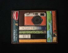 Load image into Gallery viewer, Image shows a brown Yoshi leather card holder. Design features appliqued leather travel-themed book spines and a vintage orange leather appliqued camera sat on top of the books.