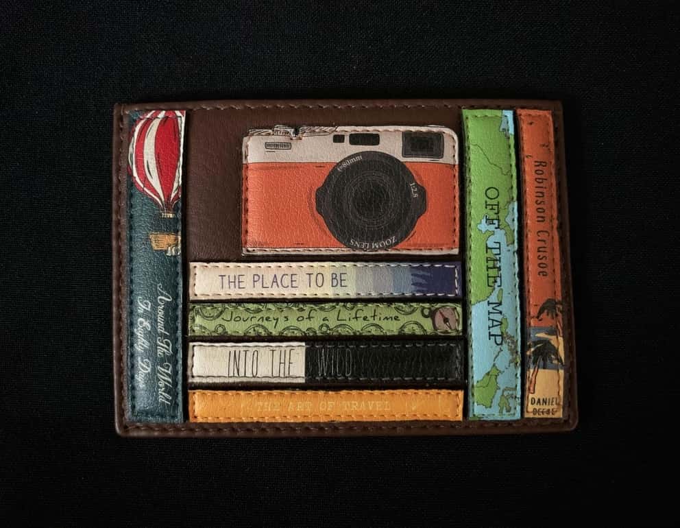 Image shows a brown Yoshi leather card holder. Design features appliqued leather travel-themed book spines and a vintage orange leather appliqued camera sat on top of the books.