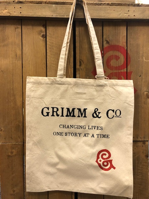 Image shows a natural beige cotton tote bag screen printed with Grimm & Co, Changing Lives One Story At A Time with the red G monogram.