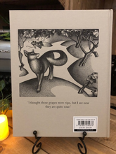 Load image into Gallery viewer, Image of the back cover of the cloth bound hardback book of Aesop&#39;s Fables in a book stand with candles