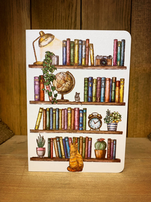 Image shows the front of the Bookshelf Card. It features a full-length illustration of shelves, which contain brightly coloured books, a lamp, a camera, a globe, a clock, plants, a mouse, and a cat sat by the bottom shelf. There is no text. The design is printed on white pearlescent card.