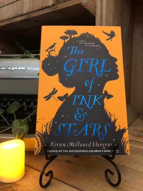 Image of the front cover of the paperback book The Girl of Ink and Stars, written by Kiran Millwood Hargrave. Displayed on a book stand with candles.
