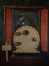 Load image into Gallery viewer, Image shows a round edition fairy door stuck onto a background of a fireplace. Accessories of window frames, decorative hinges and door knocker, and a standing sign are also included in the image.