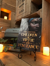 Load image into Gallery viewer, Image of front cover of paperback book Children of Vengeance and Virtue stood in book stand with a candle