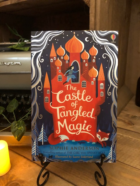 Image of the front cover of the paperback book The Castle of Tangled Magic, written by Sophie Anderson and illustrated by Saara Soderlund. Displayed on a book stand with candles.
