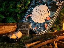 Load image into Gallery viewer, Image showing an outdoor themed setting with fake grass and living plant wall. In the foreground is a log and some firewood kindling lit as though with the glow of firelight and a Celtic Tales book displayed with the Folktales by the Fireside Scent Portal and a mini bowl containing two wax melt cubes.