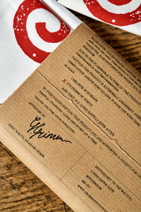 Image shows the back of the kraft paper label of the Giant Handkerchief, otherwise known as a cotton tea towel. Label lists the variety of slogans available to purchase.