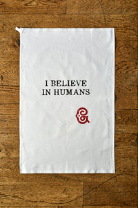 Image shows the Giant Handkerchief unfolded to display slogan printed in black on white cotton tea towel. Slogan reads 'I BELIEVE IN HUMANS' with the Grimm & Co red 'G' monogram underneath.