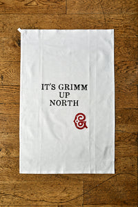 Image shows the Giant Handkerchief unfolded to display slogan printed in black on white cotton tea towel. Slogan reads 'IT'S GRIMM UP NORTH' with the Grimm & Co red 'G' monogram underneath.